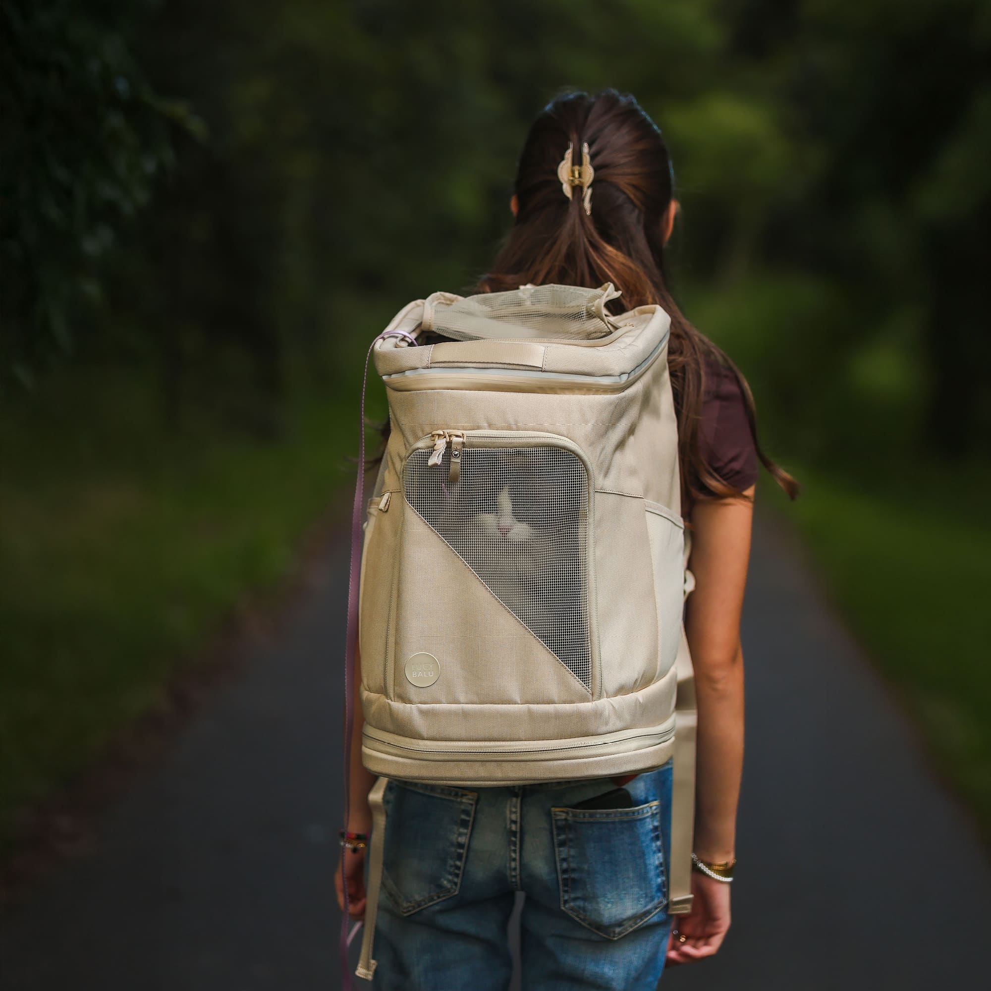 HOP-ON Katzenrucksack in Sand, wird von einer Frau auf einem Waldweg getragen, Katze schaut durch das Netzfenster